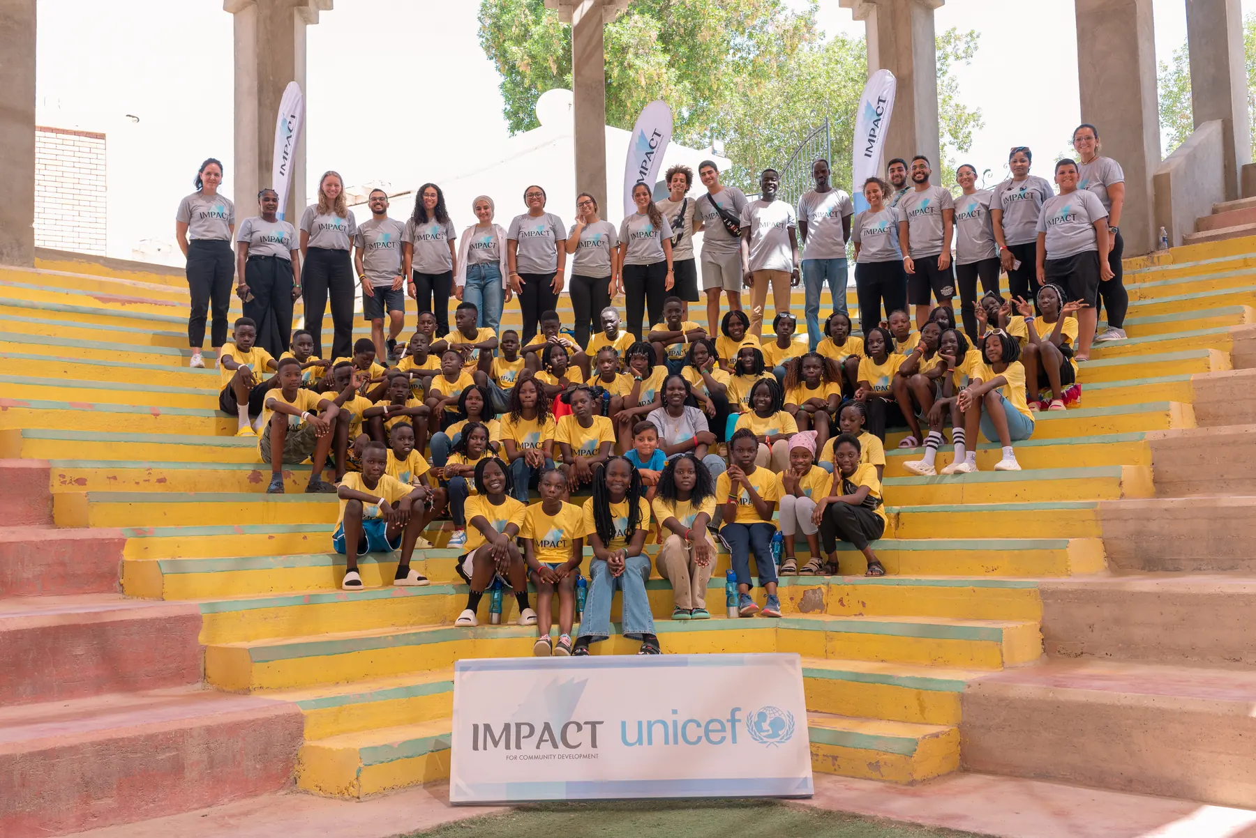 Students and facilitators of the Mazaji camp gathered on outdoor steps in front of an Impact and UNICEF banner